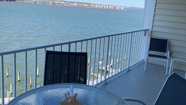 Ein Balkon mit Glastisch und Stühlen, mit Blick auf ein Gewässer und eine entfernte Küstenlinie.