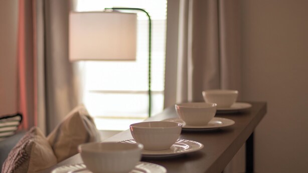 Une table à manger avec des bols et des assiettes blancs, une lampe et une fenêtre avec des rideaux.