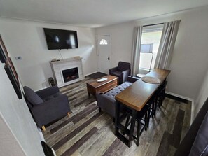 A modern living room with a fireplace, a flat-screen TV, a dining area with a long table and bar stools, and a sofa.