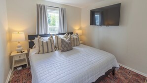 Une chambre à coucher bien aménagée, avec un grand lit, deux tables de chevet avec des lampes, un téléviseur à écran plat fixé au mur et une fenêtre avec des rideaux.