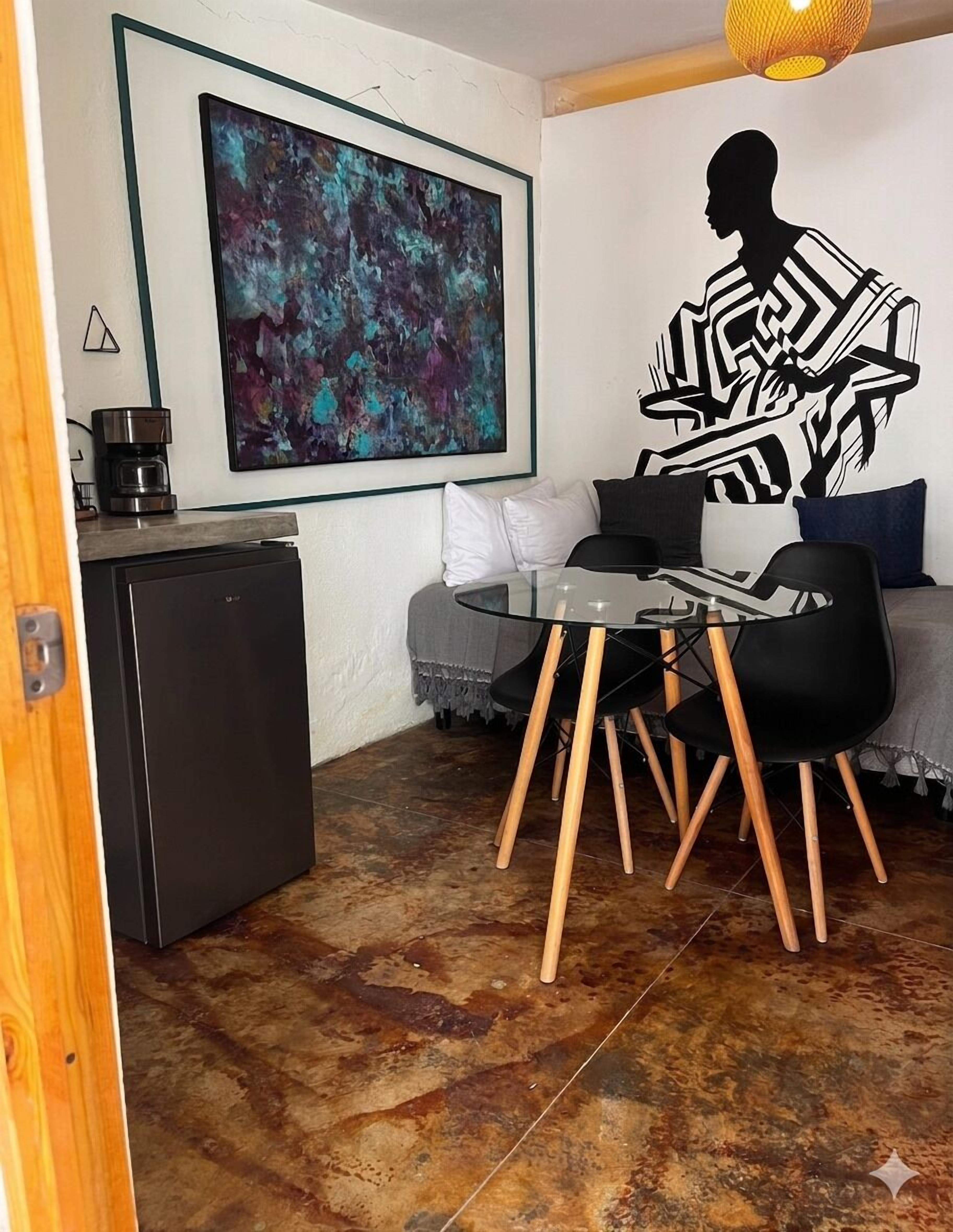 A compact living space with a kitchenette, a dining area with a glass table and chairs, a sofa, and a large abstract painting on the wall.