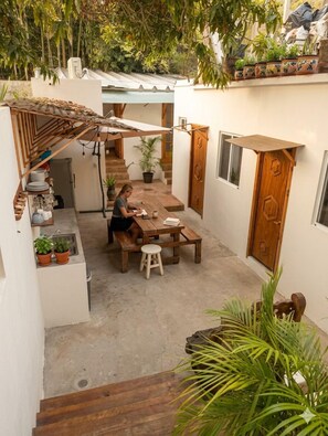 Un patio con una mesa y sillas de madera, una persona escribiendo, plantas en macetas y una escalera que conduce a un nivel superior.