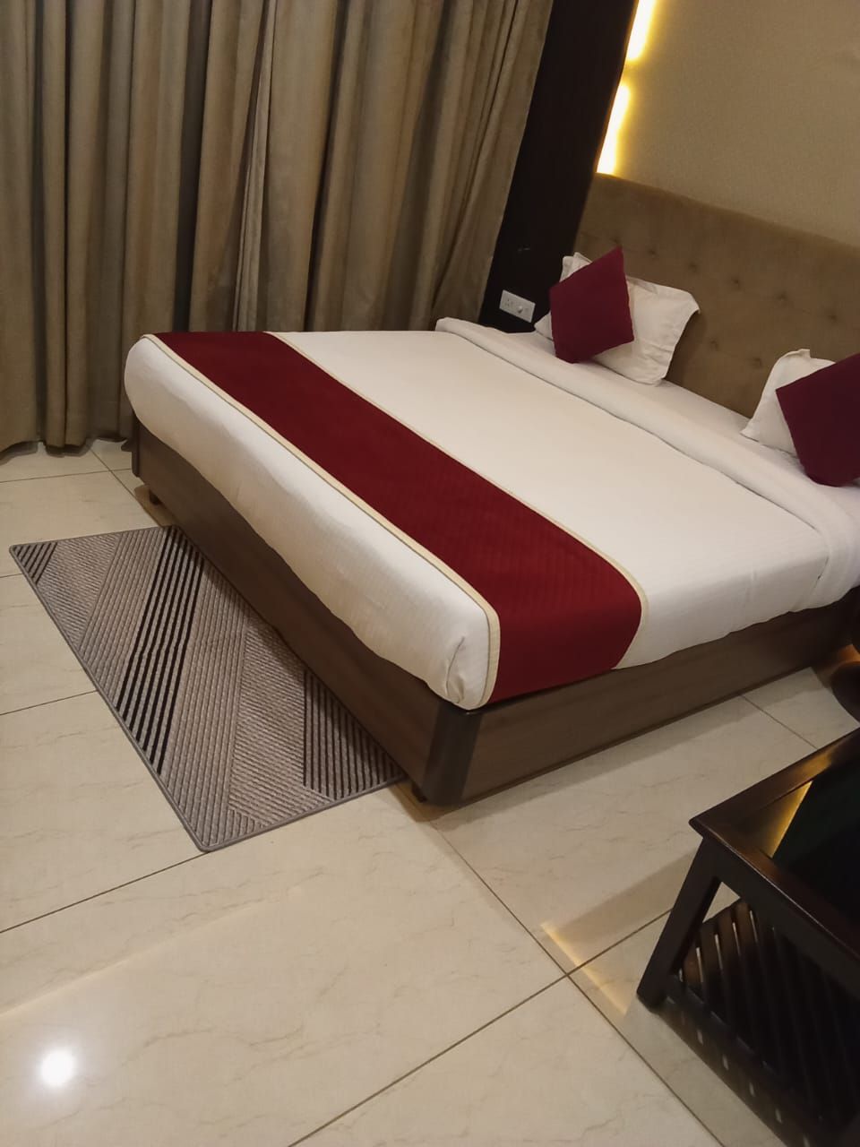 A hotel room with a large bed, beige curtains, a red and white bedspread, and a patterned rug.