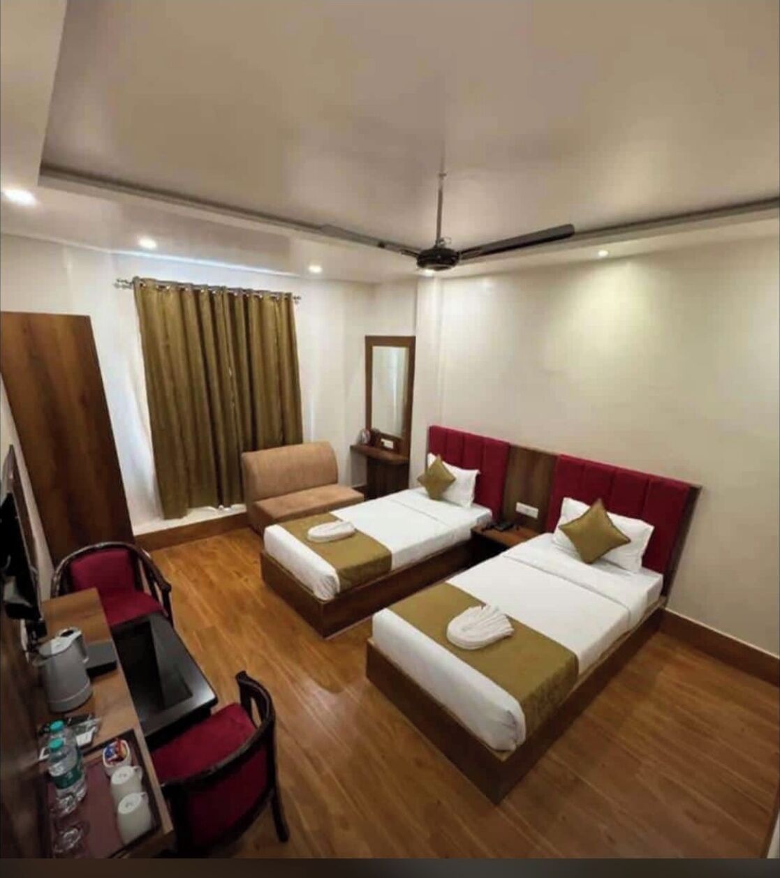 A hotel room with two single beds, a desk with a chair, a small table with a kettle, and a mirror on the wall.