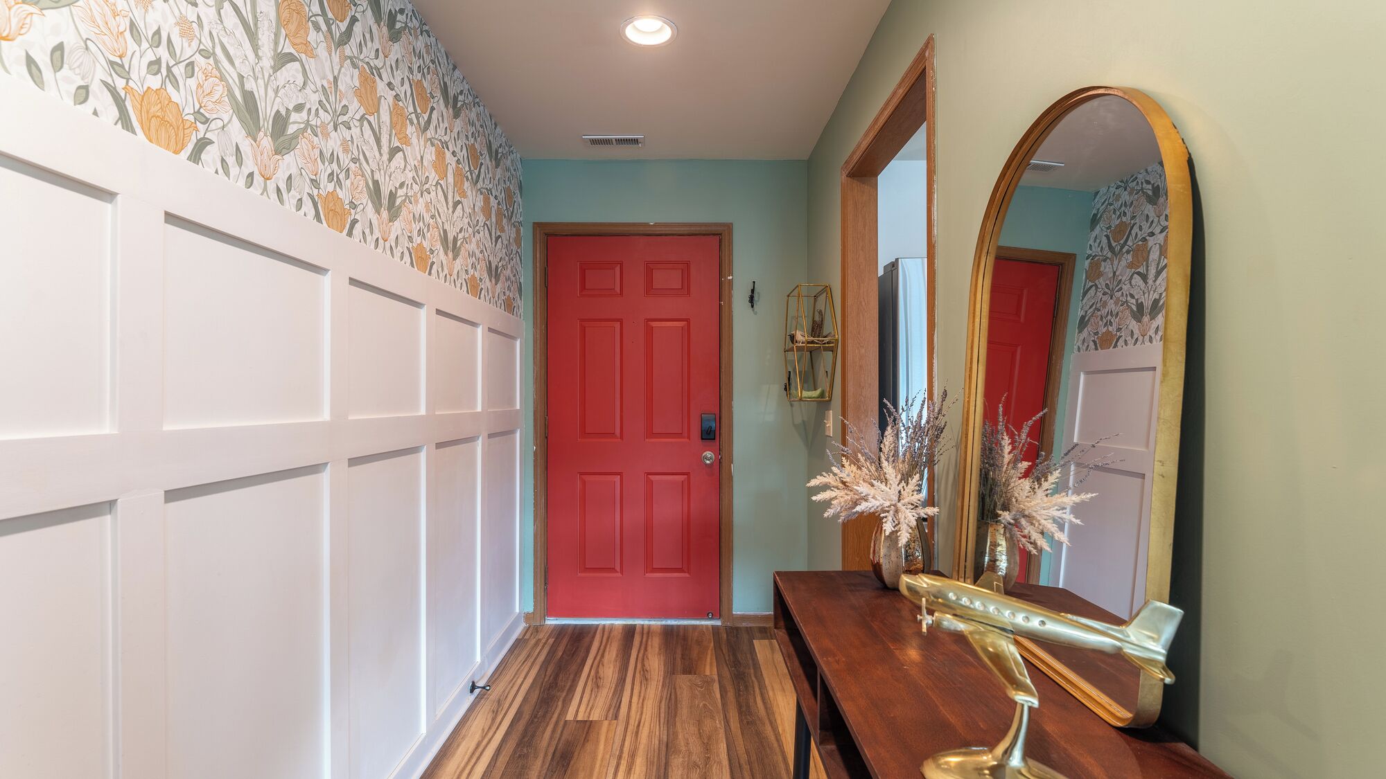A hallway with a red door, a wooden bench, a mirror, and a decorative wall with floral wallpaper.