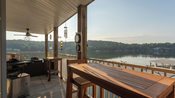 A covered outdoor deck with a wooden table and chairs, a grill, and a view of a lake with a forested shoreline.