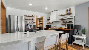 A modern kitchen with a central island, stainless steel appliances, wooden shelves, and a small dining area with chairs.