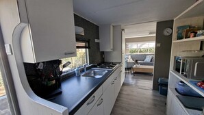 Een kleine keuken met een spoelbak, fornuis en kastjes, die uitkomt op een zithoek met een bank en een raam.