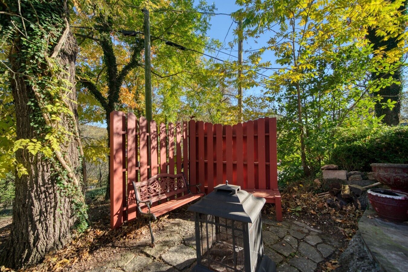 Un conjunto de banco y mesa de madera frente a una valla roja en un jardín con árboles y pavimento de piedra.