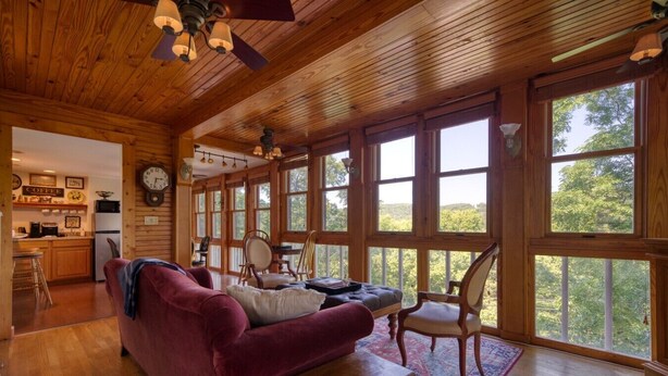 Una habitación amplia con techo de madera, ventanas grandes, un sofá rojo, una mesa de madera y una cocina visible a través de una puerta abierta.