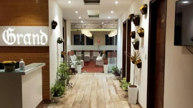 Zona de recepción de hotel con un letrero que dice 'Grand', un mostrador de recepción, plantas en macetas y vista al interior.