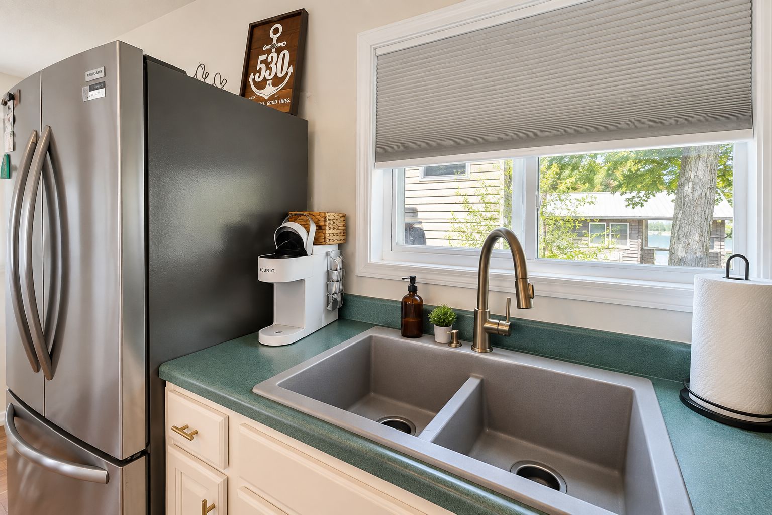 A kitchen with a stainless steel refrigerator, a Keurig coffee maker, a sink, and a window with blinds.
