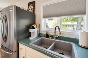 Una cocina con refrigerador de acero inoxidable, cafetera Keurig, un lavamanos y un ventanal con persianas.