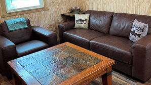 A cozy living room with a brown leather sofa, a wooden coffee table, and a window with a view of a lake. - Bass Lodge - Lakefront - New (Commerce)