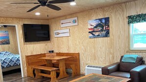A cozy living room with wooden flooring, a ceiling fan, a TV mounted on the wall, a wooden coffee table, a sofa, and a painting on the wall. - Bass Lodge - Lakefront - New (Commerce)