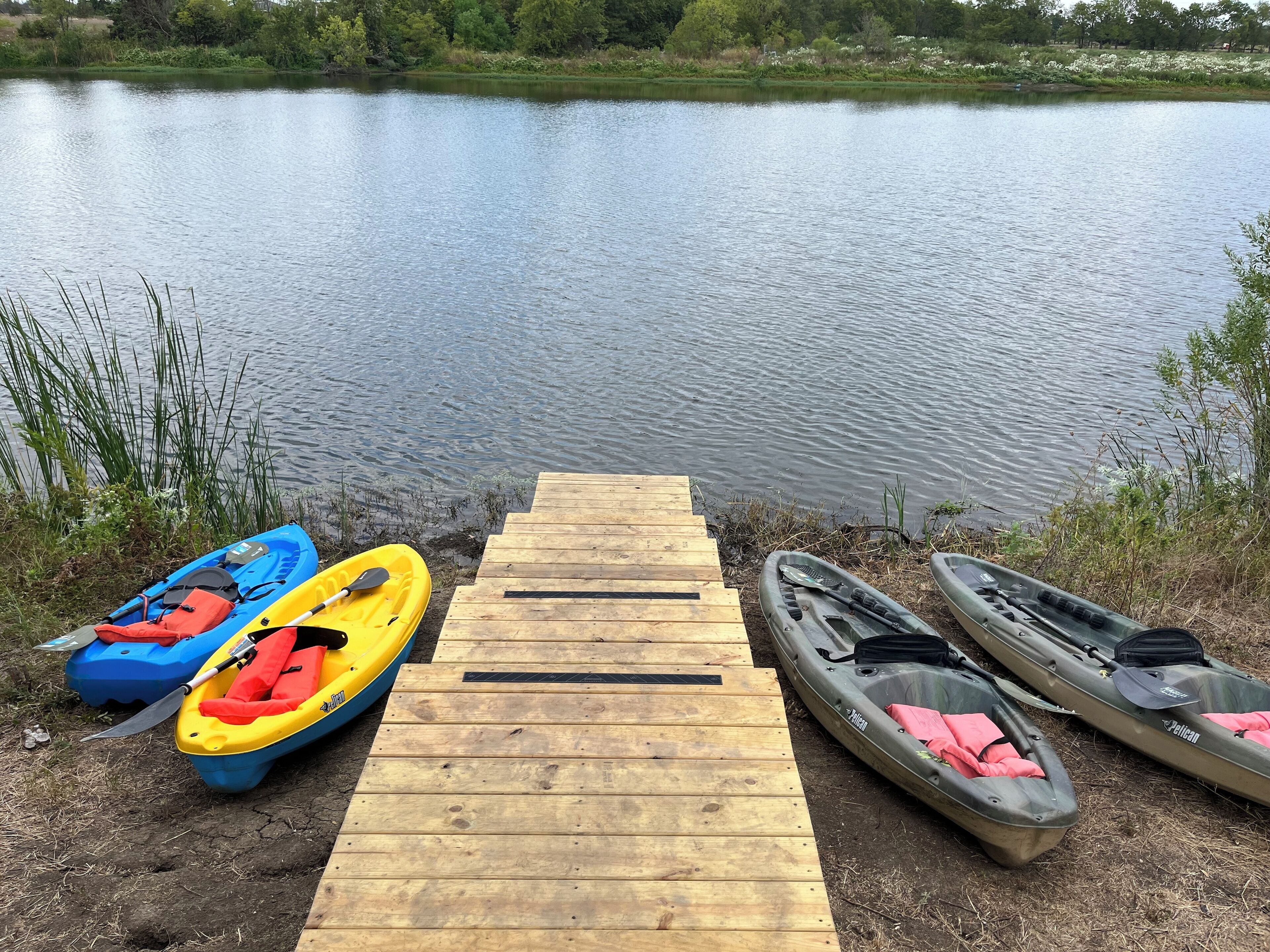 Un muelle de madera que se adentra en un lago tranquilo, con kayaks y una canoa alineados en la orilla.
