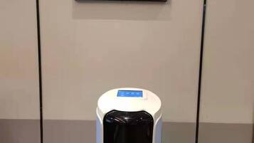 Un objeto cilíndrico blanco con una pantalla digital en la parte superior, posiblemente un equipo o quiosco, frente a una pared lisa con un cuadro colgado encima.