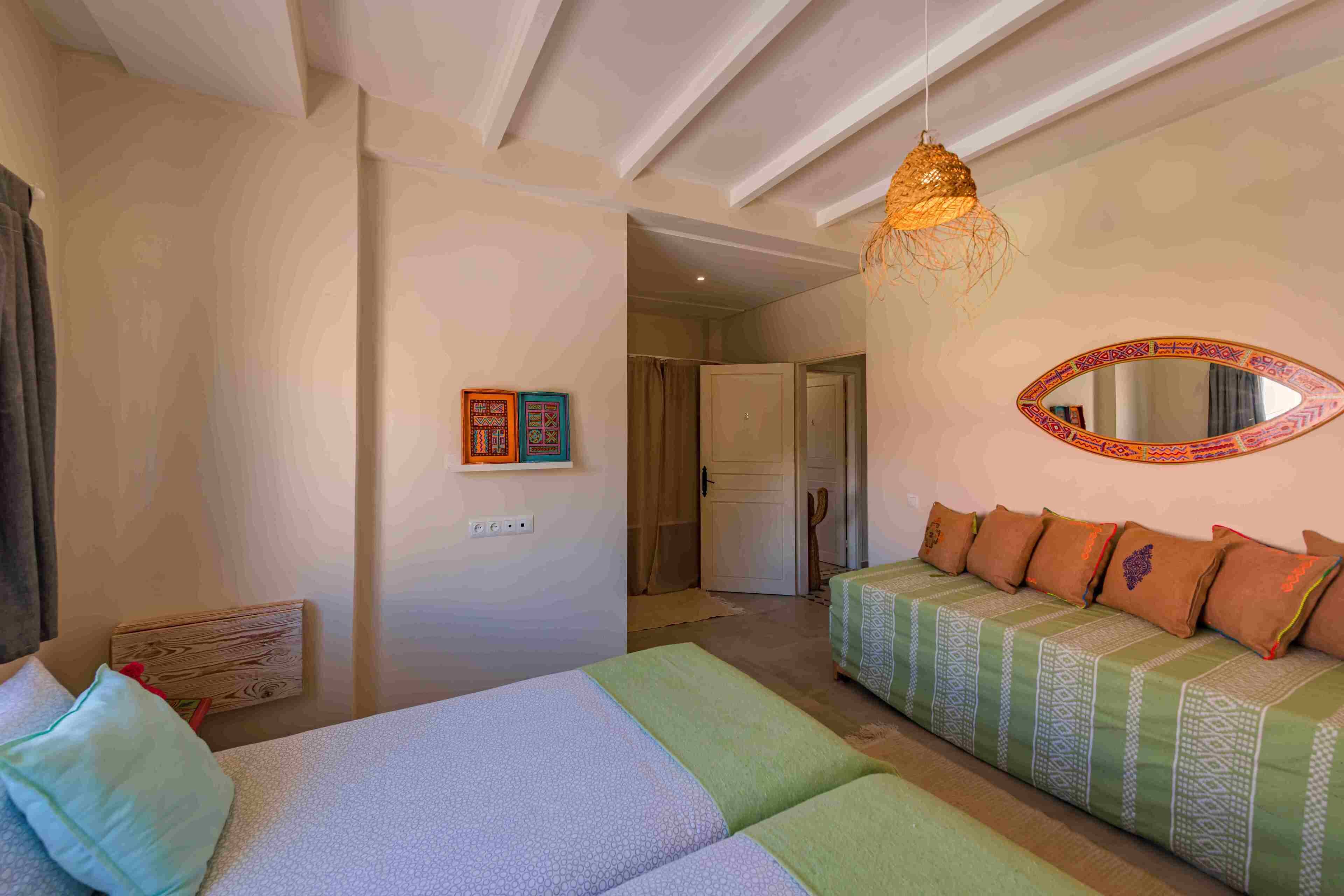 A room with a bed, a sofa, a hanging light fixture, a mirror, and a door leading to another room.