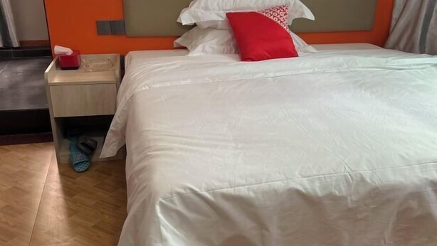 Habitación de hotel con una cama, una mesita de noche, un almohadón rojo, una caja de pañuelos y pantuflas.