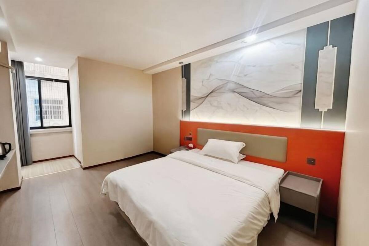 A hotel room with a large bed, a nightstand, a wall-mounted light, and a window with curtains.