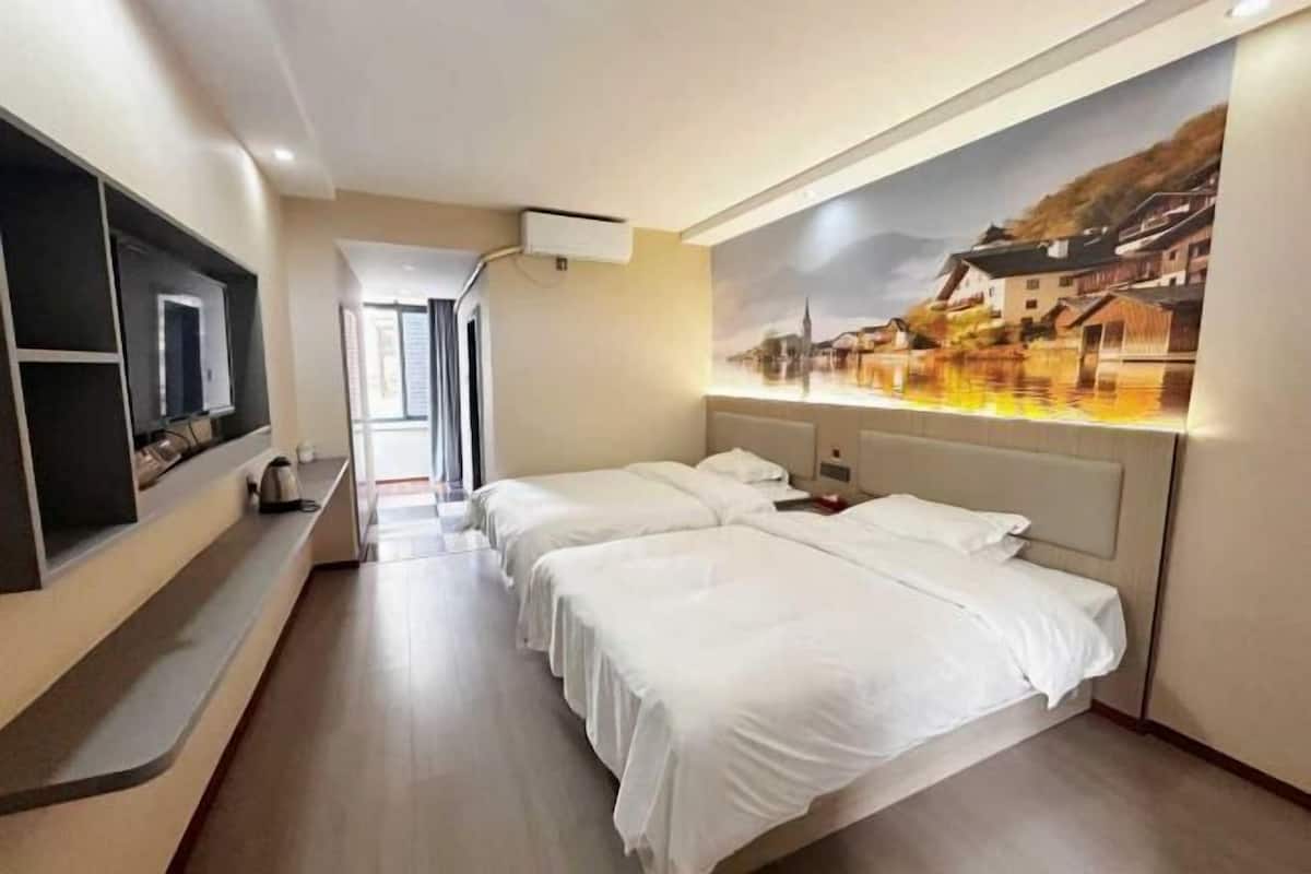 A hotel room with two beds, a flat-screen TV, a painting on the wall, and a view of a town through the window.