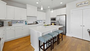 A modern kitchen with white cabinets, a central island with bar stools, stainless steel appliances, and a 'gather here' sign.