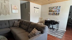 A modern living room with a gray sectional sofa, a small round table, a ceiling fan, and a dining area with a black chair and a small table. - Riverside Retreat 2 ~ One Story ~ 3bd/2ba ~ Steps to the River & Main St (Madison)