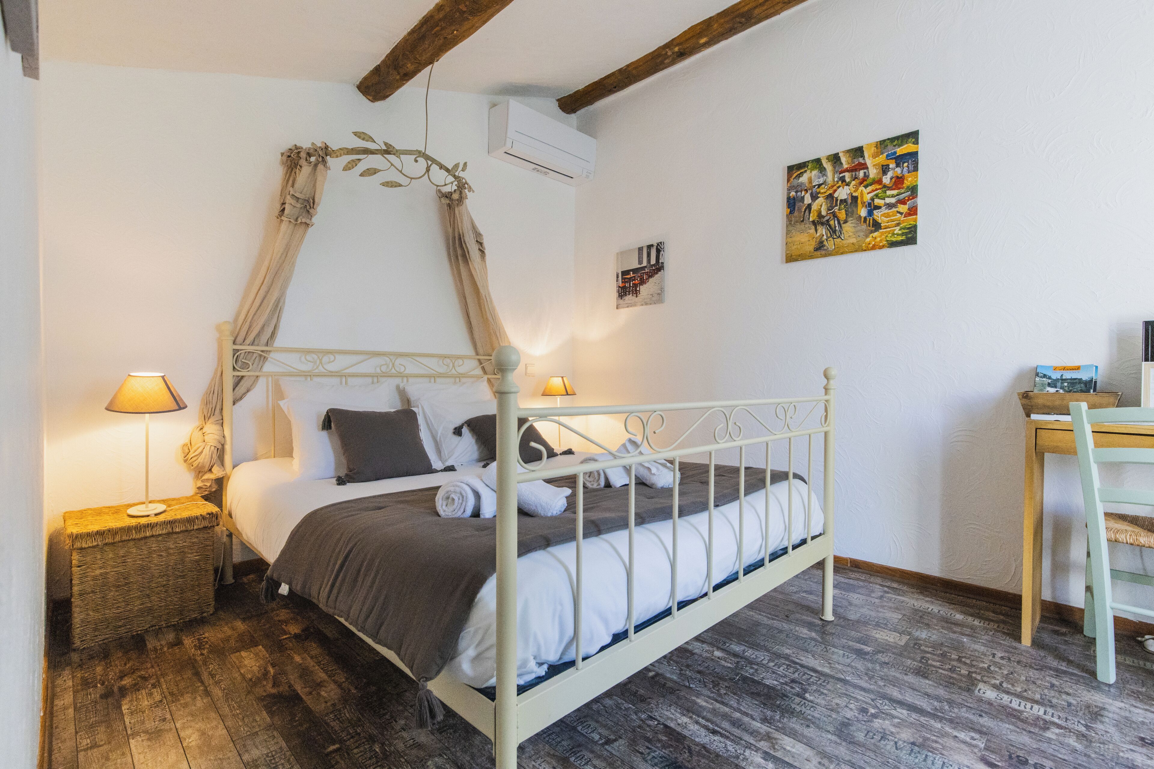 A bedroom with a four-poster bed, a bedside table with a lamp, a wooden floor, and a desk with a chair.