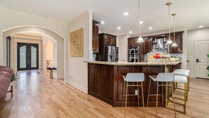 Una cocina moderna con armarios de madera, electrodomésticos de acero inoxidable y encimera de mármol. Hay tres taburetes de bar y luces colgantes sobre la encimera.