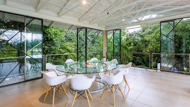 A modern dining area with a glass table and white chairs, surrounded by large windows offering a view of lush greenery. - Casa Paraíso, Valle de Bravo. (Valle de Bravo)