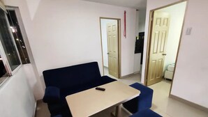 Ein kleines Zimmer mit einem blauen Sofa, einem kleinen Tisch und zwei blauen Stühlen. Links ist ein Fenster und rechts eine Tür.