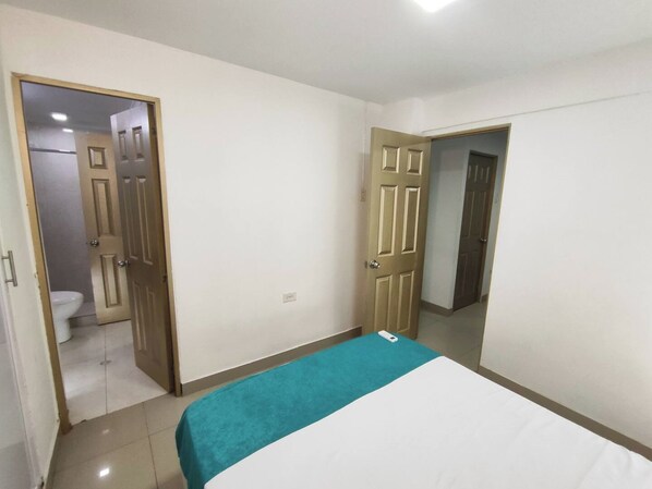 Un dormitorio con cama, un baño visible a través de una puerta abierta y un pasillo que conduce a otra habitación.