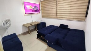 Ein Zimmer mit dunkelblauer Couch, einem kleinen Tisch, einem an der Wand montierten Fernseher und einem Fenster mit Jalousien.