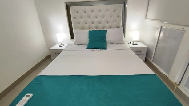 Ein Hotelzimmer mit einem Bett, zwei Nachttischen mit Lampen, einer türkisblauen Tagesdecke und einer Fernbedienung auf dem Bett.