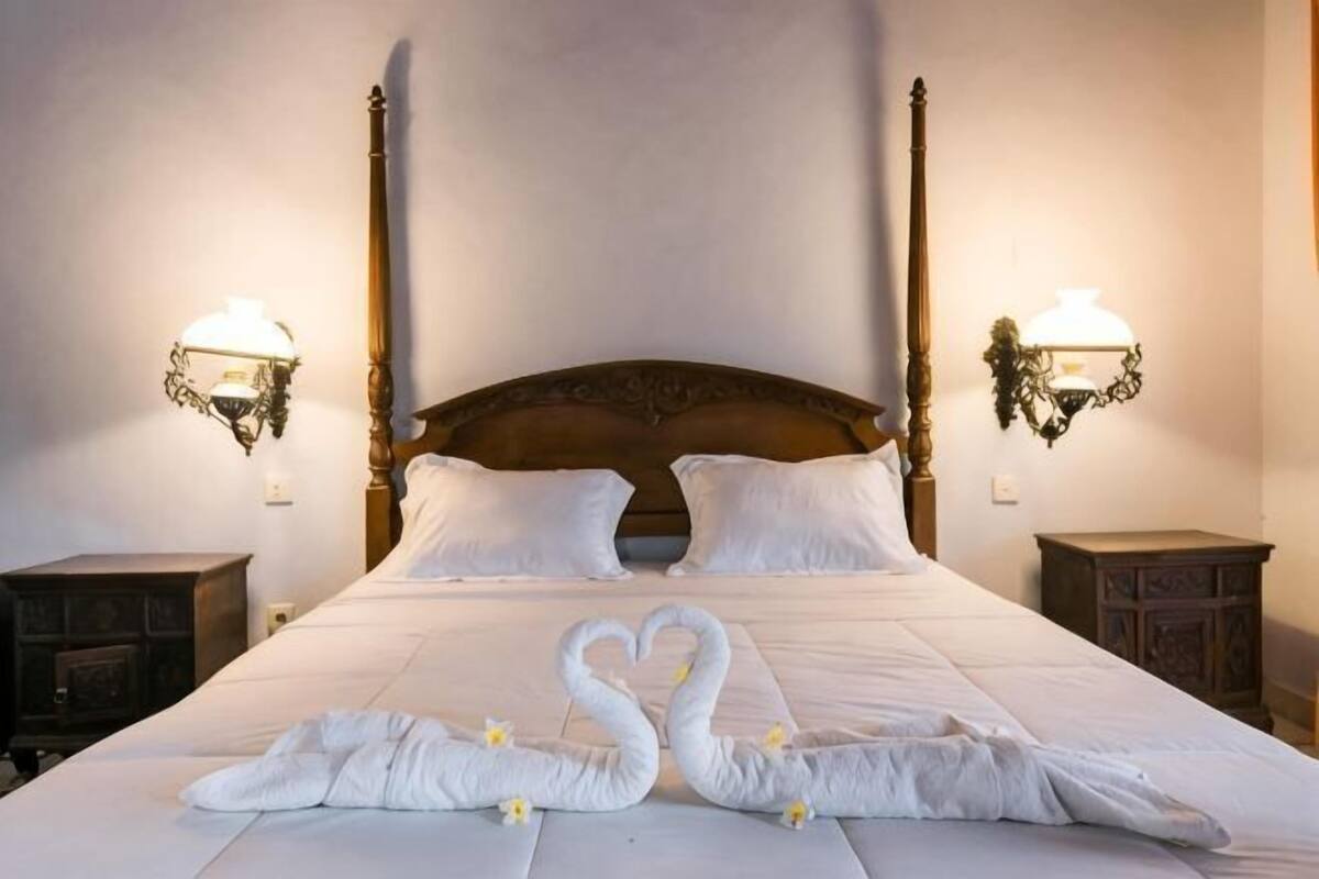 Una cama con cabecera de madera, dos lámparas fijadas a la pared y un arreglo decorativo con cisnes sobre el edredón.