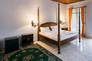 Una cama con dosel con sábanas blancas, dos lámparas fijadas a la pared, una mesita de noche, un tapete verde estampado y una ventana con cortinas.