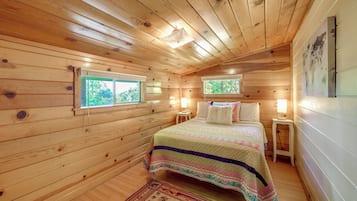 Une chambre confortable dans un chalet, avec un plafond en bois, deux fenêtres, un lit recouvert d’une courtepointe colorée, des tables de chevet avec des lampes et un tapis sur le sol.