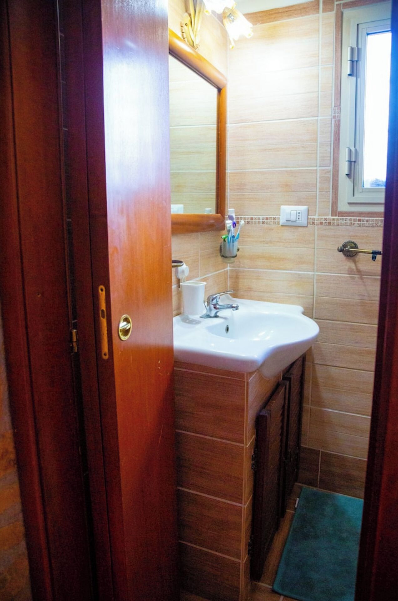 Een badkamer met een houten deur, een wit wastafelmeubel, een spiegel en een raam.