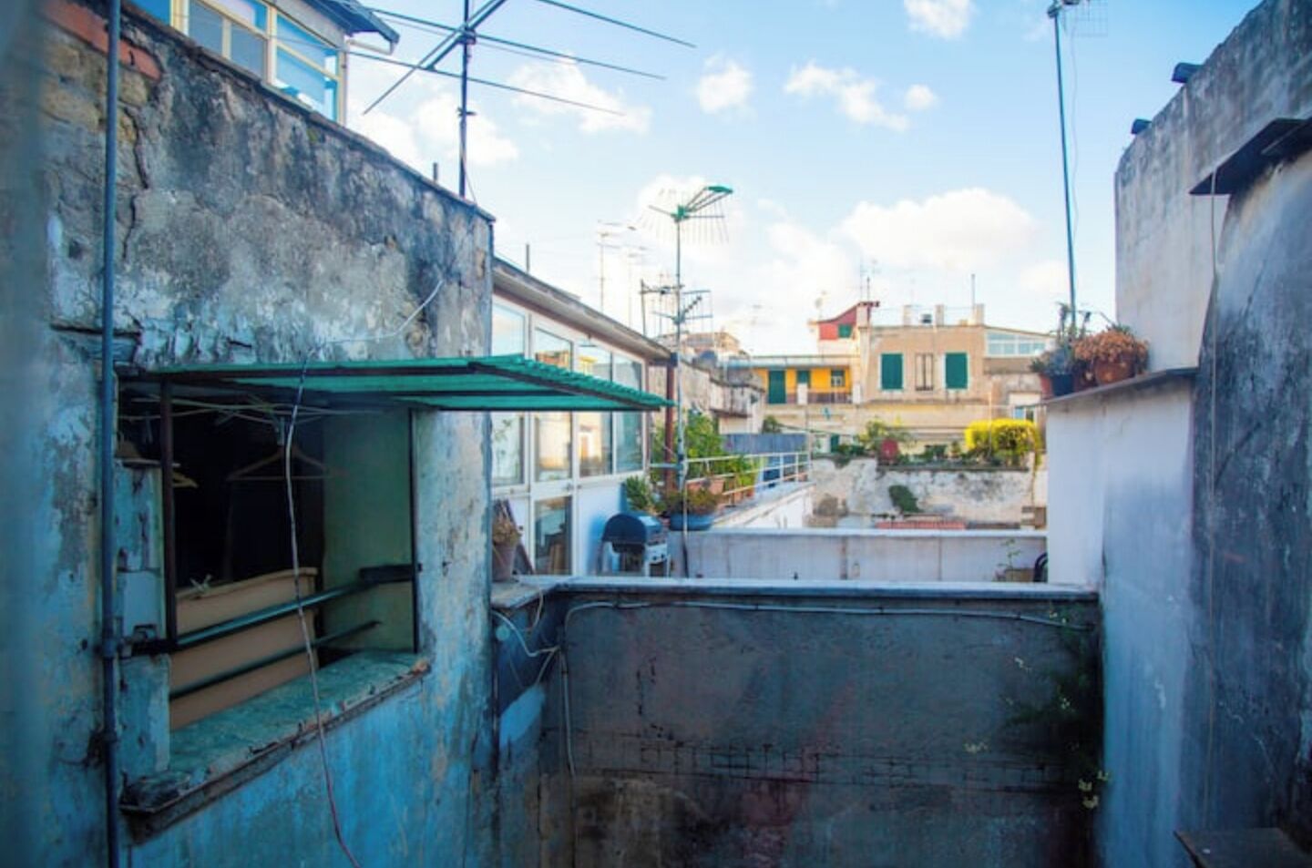 A narrow alleyway with a small, open-air structure on the left, a metal barrier in the center, and buildings in the background.