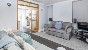 A modern living room with a grey sofa, a coffee table, and a TV stand. There is a kitchen visible through an open door. - 4 bedroom accommodation in Gwithian (Gwithian)