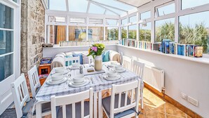 A sunlit dining area with a table set for four, surrounded by white chairs, a white radiator, and a bookshelf filled with books. - 4 bedroom accommodation in Gwithian (Gwithian)