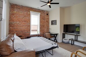 Quarto com parede de tijolos, cama, escrivaninha, cadeira e televisão.
