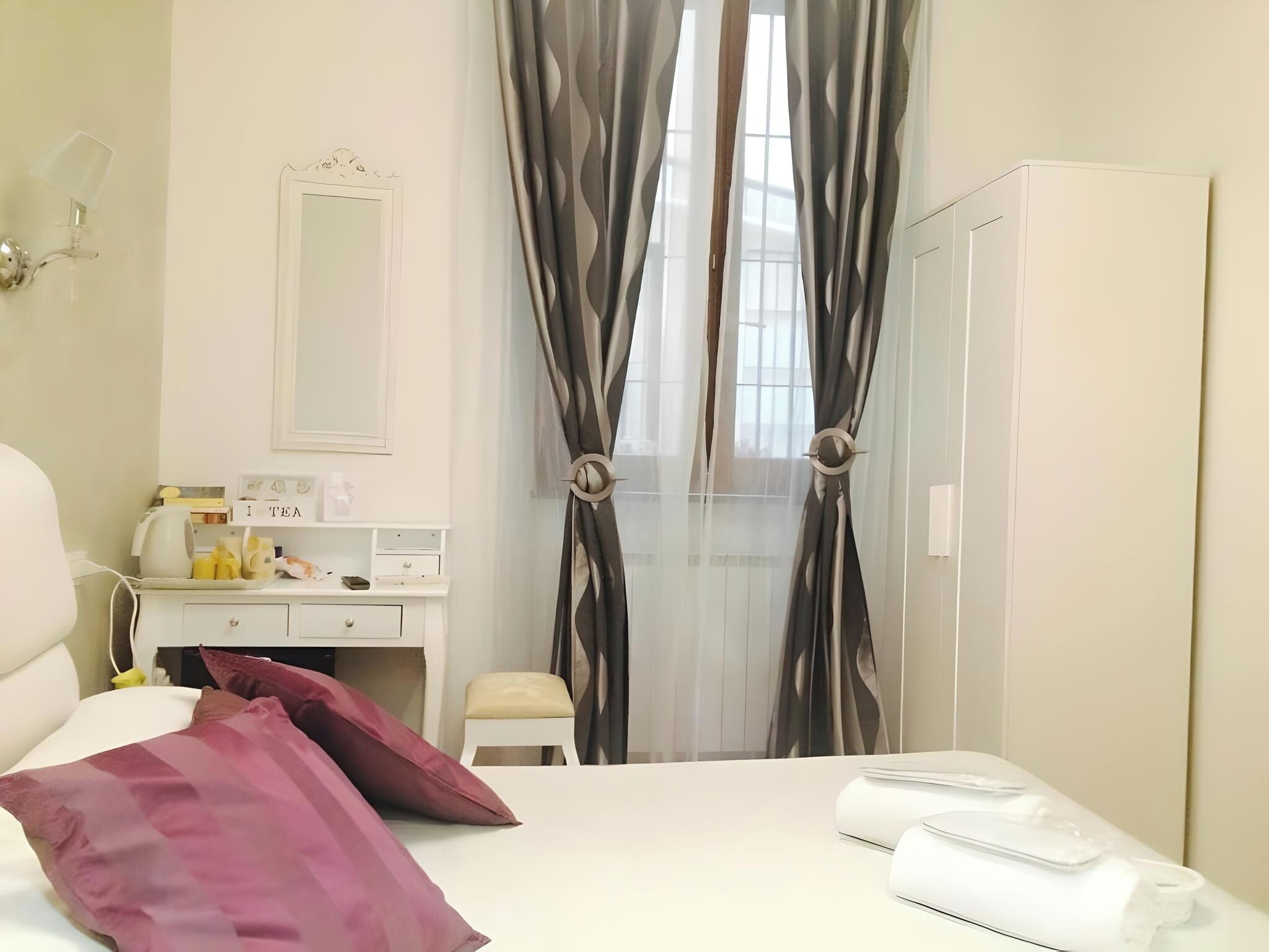A hotel room with a bed, a bedside table with a lamp, a small desk with a mirror, a chair, and a wardrobe.