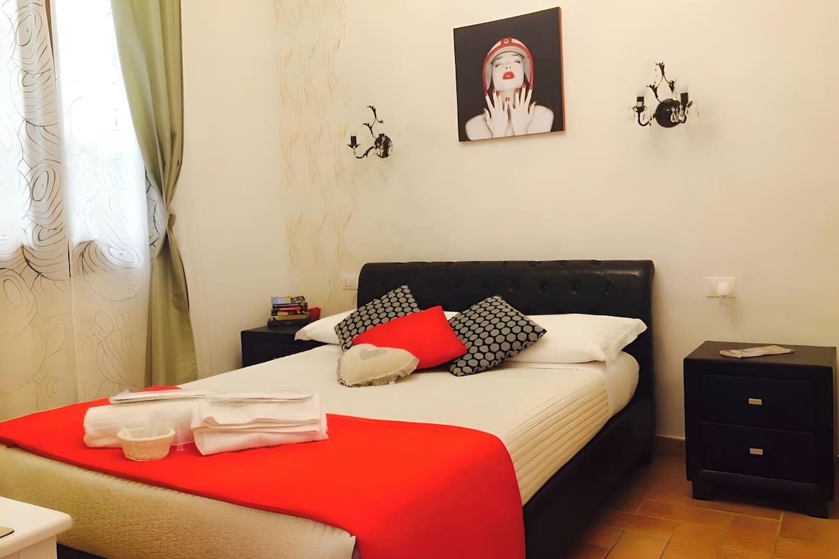 A neatly made bed with a red and white bedspread, two black nightstands with books and a candle, a wall-mounted artwork, and a window with green curtains.