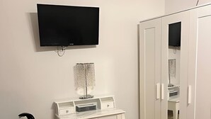 Una habitación con un escritorio blanco, una cafetera negra, un televisor en la pared y un armario con puertas espejadas.