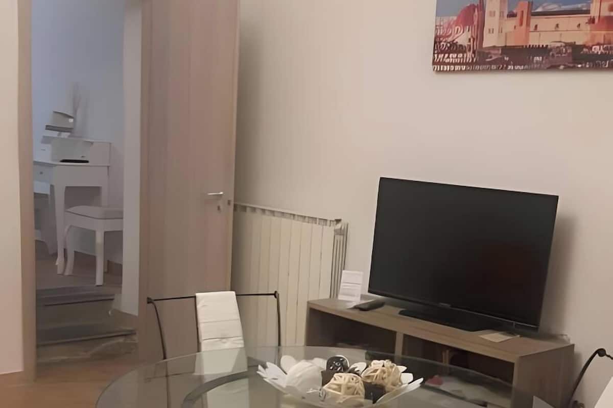 A dining area with a glass table surrounded by chairs, a television on a stand, and a painting on the wall.