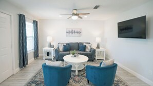 Una sala de estar con un sofá gris, dos sillones azules, una mesa de centro blanca y un ventilador de techo.