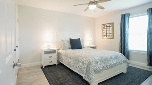 Un dormitorio con cama, dos mesitas de noche con lámparas, ventilador de techo, ventana con cortinas y una obra de arte enmarcada en la pared.