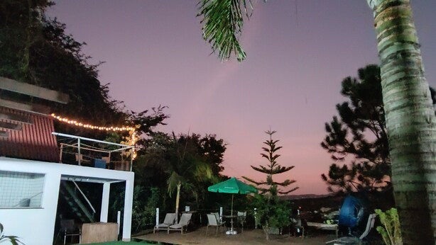 A swimming pool with lounge chairs and umbrellas, surrounded by trees and a building with string lights. - Aqui no Camping (Brumadinho)