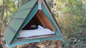 A small, triangular wooden tent with a green roof, a sleeping area with a white mattress, and a decorative hanging ornament. - Aqui no Camping (Brumadinho)